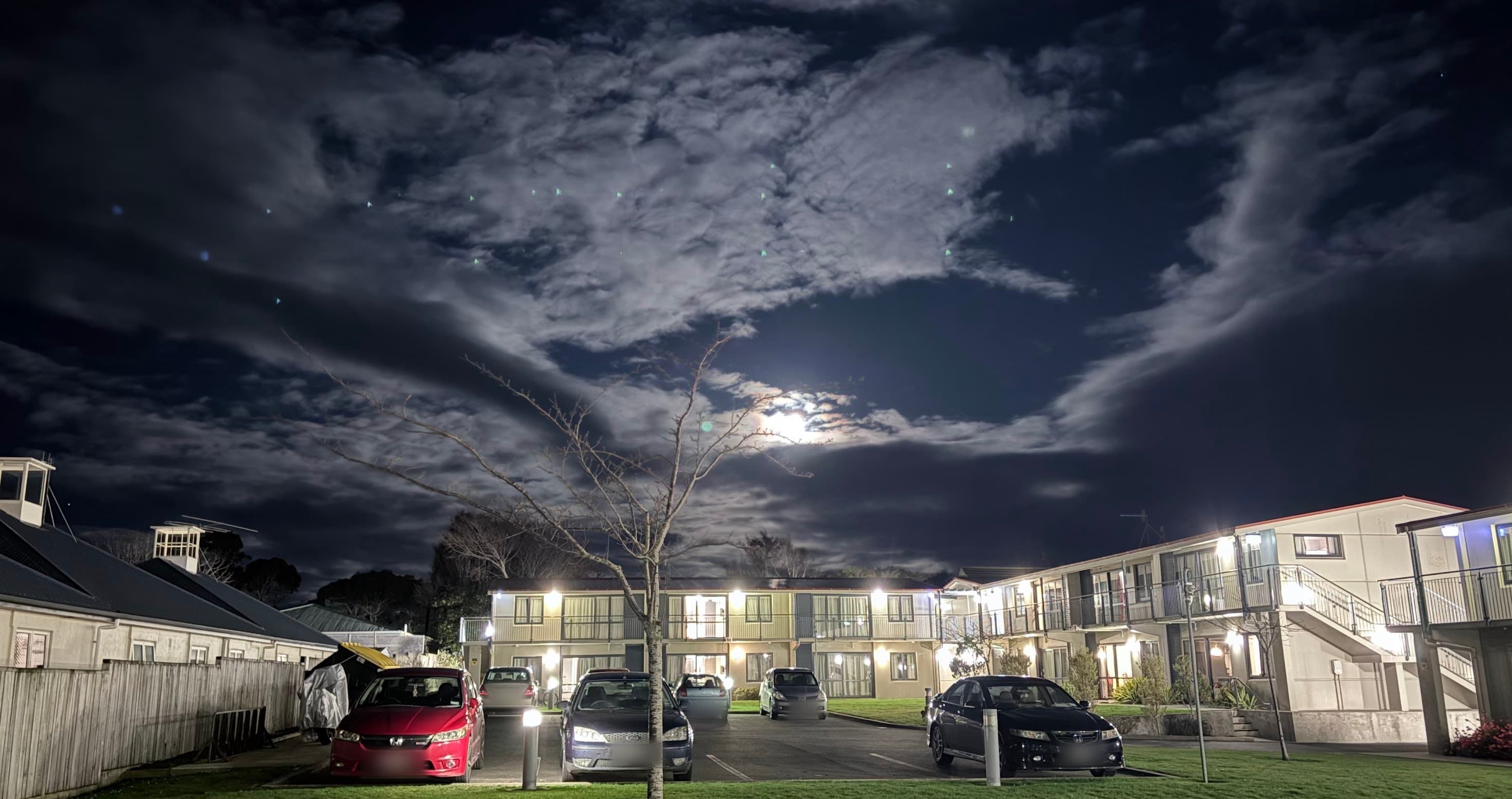 A car park at night outside two-storey accommodation blocks, balcony lights warm against the dark, with the moon breaking through heavy cloud above a bare young tree